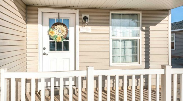 Porch of a house with beige vinyl wood siding and railing. There is a white enrty door with wreath on top of the glass panels near the single hung window with curtain